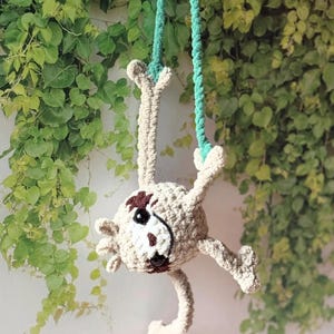 May include: A crocheted sloth toy in light beige yarn with dark brown eye patches and a stitched nose. The sloth has black plastic eyes and is suspended by two teal green braided cords attached to its front paws, hanging in a downward position.
