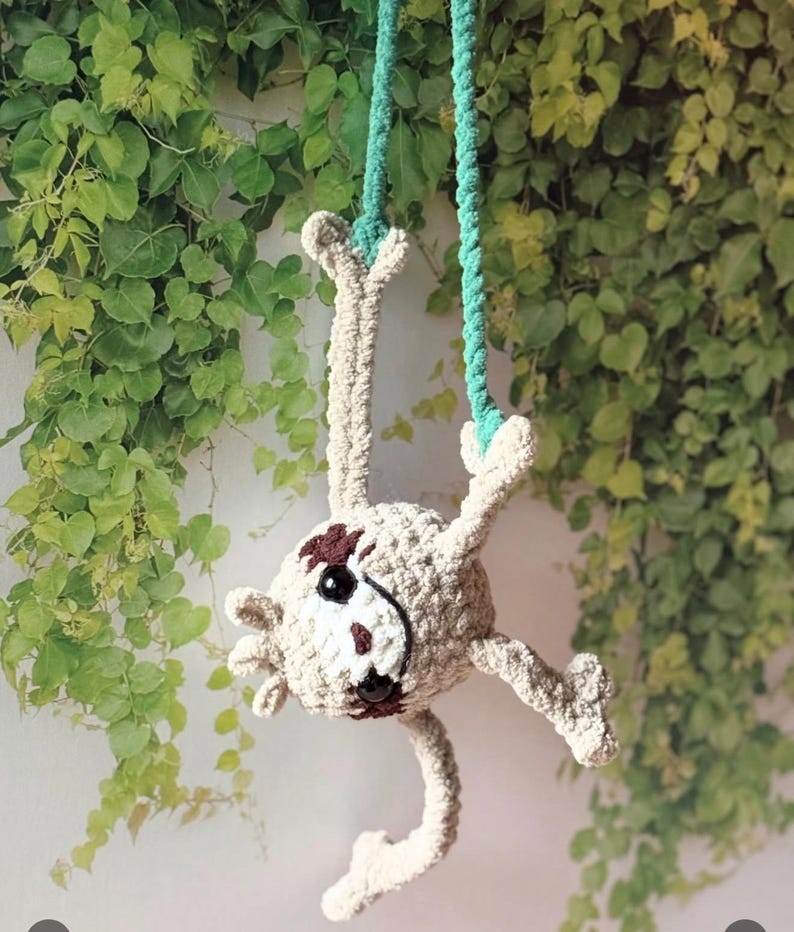 May include: A crocheted sloth toy in light beige yarn with dark brown eye patches and a stitched nose. The sloth has black plastic eyes and is suspended by two teal green braided cords attached to its front paws, hanging in a downward position.