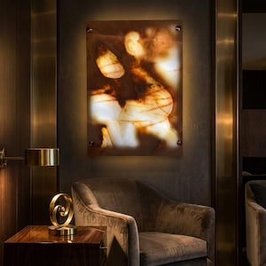 May include: A backlit, rectangular wall art piece with a natural stone appearance, featuring shades of brown, white, and amber. The artwork is mounted on a dark wall with visible mounting hardware. A gold-toned lamp and decorative sculpture sit on a wooden side table.