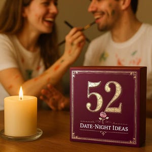 May include: A lit ivory candle sits on a glass plate next to a burgundy box with gold lettering. The box reads "52 DATE-NIGHT IDEAS" with a rose graphic. Two people are in the background, smiling and painting each other's faces.