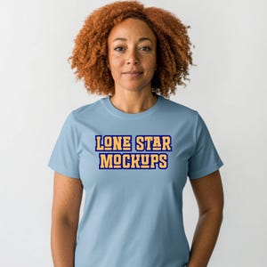 May include: A light blue t-shirt with the text "LONE STAR MOCKUPS" in a bold, two-tone design. The shirt has short sleeves and a crew neck. The model has curly auburn hair and is facing the camera.
