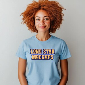 May include: A light blue t-shirt with the text "LONE STAR MOCKUPS" in a yellow and blue block letter design. The shirt is short-sleeved and has a crew neck. The model has red curly hair and is smiling.