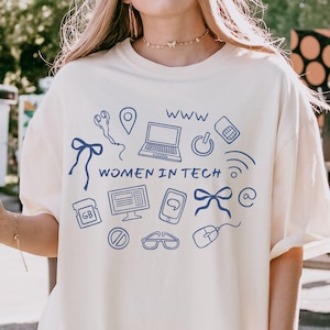 May include: Cream-colored t-shirt with a blue graphic design featuring tech-related icons and the text "WOMEN IN TECH" and "WWW". The shirt has short sleeves and a relaxed fit.