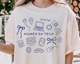 May include: Cream-colored t-shirt with a blue graphic design featuring tech-related icons and the text "WOMEN IN TECH" and "WWW". The shirt has short sleeves and a relaxed fit.