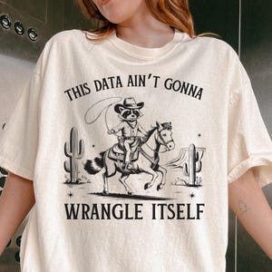 May include: Ivory-colored t-shirt with a graphic of a raccoon riding a horse, roping a lasso, and the text "THIS DATA AIN'T GONNA WRANGLE ITSELF." The design includes cacti and stars. The shirt is a relaxed fit.
