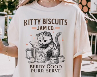 May include: Cream-colored t-shirt with the text "KITTY BISCUITS JAM CO." and a graphic of a cat stirring jam. The graphic also includes jars of jam and the words "BERRY GOOD PURR-SERVE."
