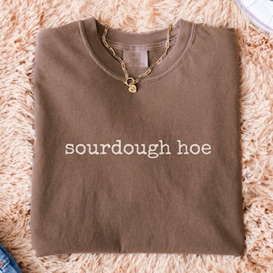 May include: Espresso-colored t-shirt with the words "sourdough hoe" in white lettering. A gold chain necklace with a pendant rests on the shirt. The shirt is on a fluffy, light brown surface, with a pair of white sneakers and blue jeans visible.