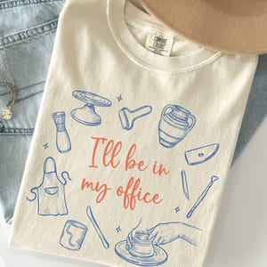 May include: Ivory-colored t-shirt with blue pottery tools and the text "I'll be in my office" in coral. The shirt is laid on top of light blue jeans and a tan hat.