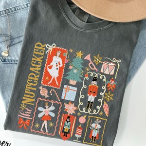 May include: A dark gray t-shirt with a colorful "The Nutcracker" graphic. The design includes illustrations of a ballerina, a nutcracker, a Christmas tree, and other festive elements. The shirt is paired with light blue jeans and a tan hat.