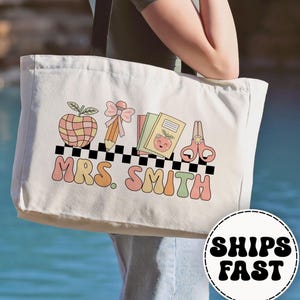 May include: Cream-colored tote bag with a retro design featuring an apple, pencil, books, scissors, and the name "MRS. SMITH" in colorful letters. The bag has a black and white checkered pattern and the words "SHIPS FAST" in a circle.