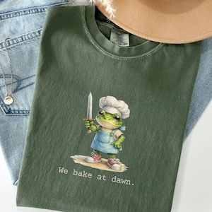 May include: Moss green t-shirt featuring a cartoon frog chef holding a knife, wearing a chef's hat and apron. The text "We bake at dawn." is printed below the frog. The shirt is paired with light blue jeans and a tan hat.