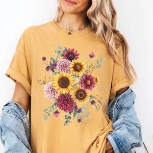 May include: Mustard yellow t-shirt featuring a floral design with sunflowers, dahlias, and other flowers in shades of yellow, pink, and purple. The shirt has short sleeves and a relaxed fit. A denim jacket is draped over the shoulders.