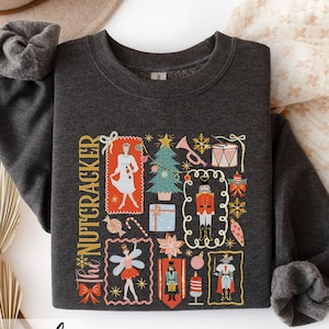 May include: Dark heather gray sweatshirt featuring a colorful graphic of The Nutcracker. The design includes illustrations of a ballerina, Christmas tree, nutcracker soldier, and other festive elements. The text "The Nutcracker" is printed vertically on the left side.
