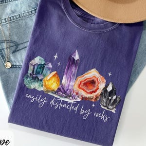 May include: A purple t-shirt with a watercolor design of colorful crystals and the text "easily distracted by rocks." The shirt is paired with light blue jeans and a tan hat.
