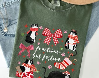 Könnte beinhalten: Ein moosgrünes T-Shirt mit einem festlichen Katzendesign. Die Grafik zeigt schwarz-weiße Katzen mit Schals und Hüten, umgeben von Schleifen, Pfotenabdrücken und einer Zuckerstange. Der Text "fractious but festive" ist ebenfalls aufgedruckt.