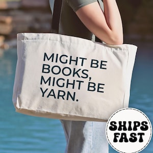 May include: A cream-colored tote bag with the text "MIGHT BE BOOKS, MIGHT BE YARN" in dark blue. The bag is held by a person. The background is a body of water and some rocks. The bag has a round sticker that says "SHIPS FAST".