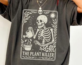 May include: Dark grey t-shirt featuring a white graphic of a skeleton watering a plant, with the text "THE PLANT KILLER" in a rectangular frame. The design includes a skull, sun, and rose. The shirt has short sleeves.