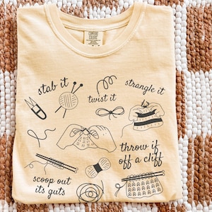 May include: A light yellow t-shirt with a crew neck and short sleeves. The shirt features black line art illustrations of knitting tools and phrases like "stab it" and "throw it off a cliff." The word "Butter" is printed in the bottom left corner.