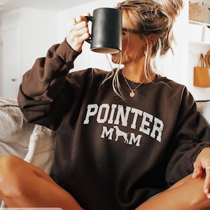 May include: Dark chocolate brown sweatshirt with the text "POINTER MOM" in white, featuring a silhouette of a dog. A black mug is held up. The words "Dark Chocolate" are at the bottom of the image.
