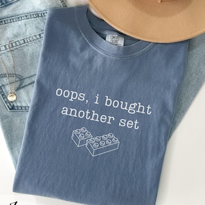 May include: A blue t-shirt with the text "oops, I bought another set" and a graphic of two toy blocks. The shirt is folded on top of a pair of blue jeans and a tan hat.
