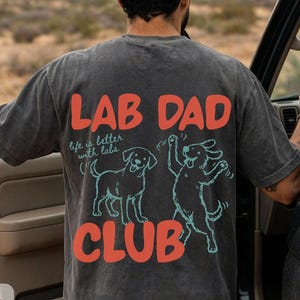 Puede incluir: Camiseta gris oscuro con el texto "LAB DAD CLUB" en rojo y blanco. La camiseta presenta ilustraciones de dos perros Labrador y la frase "life is better with labs".