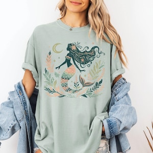 May include: A light green t-shirt featuring a mermaid graphic. The mermaid has long flowing hair, a floral crown, and a patterned tail. The design includes a crescent moon and floral elements in shades of green, pink, and peach.