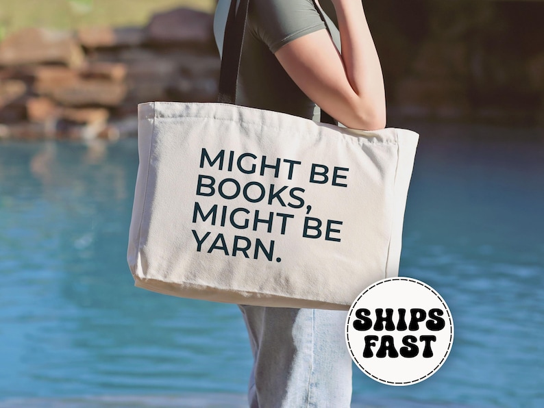 May include: A cream-colored tote bag with the text "MIGHT BE BOOKS, MIGHT BE YARN" in dark blue. The bag is held by a person. The background is a body of water and some rocks. The bag has a round sticker that says "SHIPS FAST".