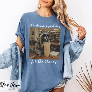 May include: A blue denim-colored t-shirt featuring a graphic of a raccoon wearing sunglasses, drinking an iced coffee, and making a peace sign in a library. The text above reads "it's always a good day" and the text below reads "for the library."