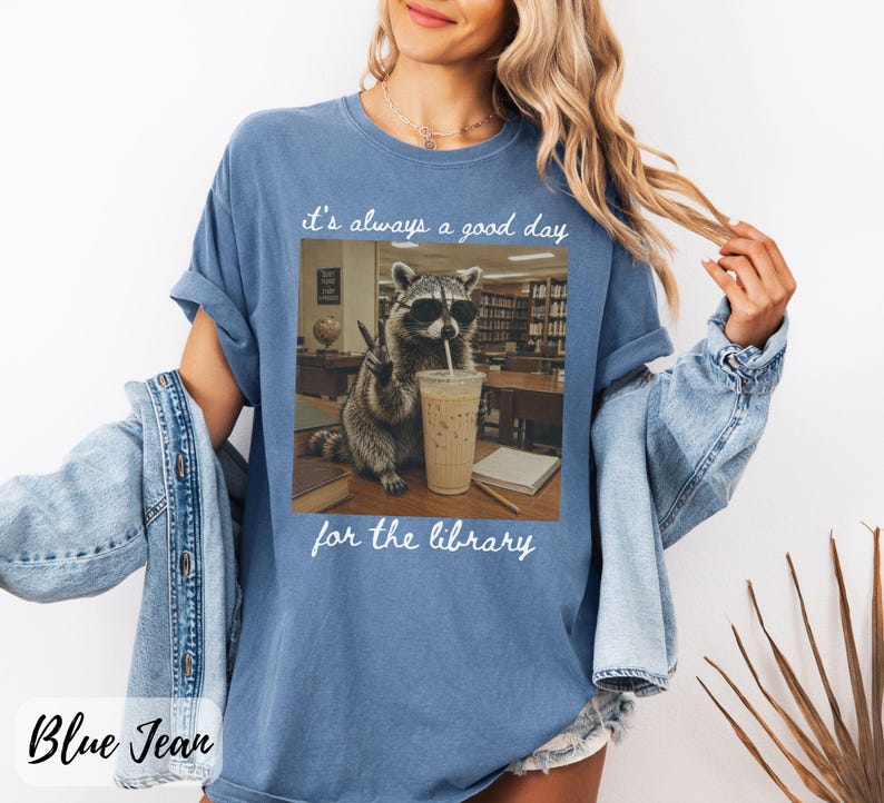 May include: A blue denim-colored t-shirt featuring a graphic of a raccoon wearing sunglasses, drinking an iced coffee, and making a peace sign in a library. The text above reads "it's always a good day" and the text below reads "for the library."