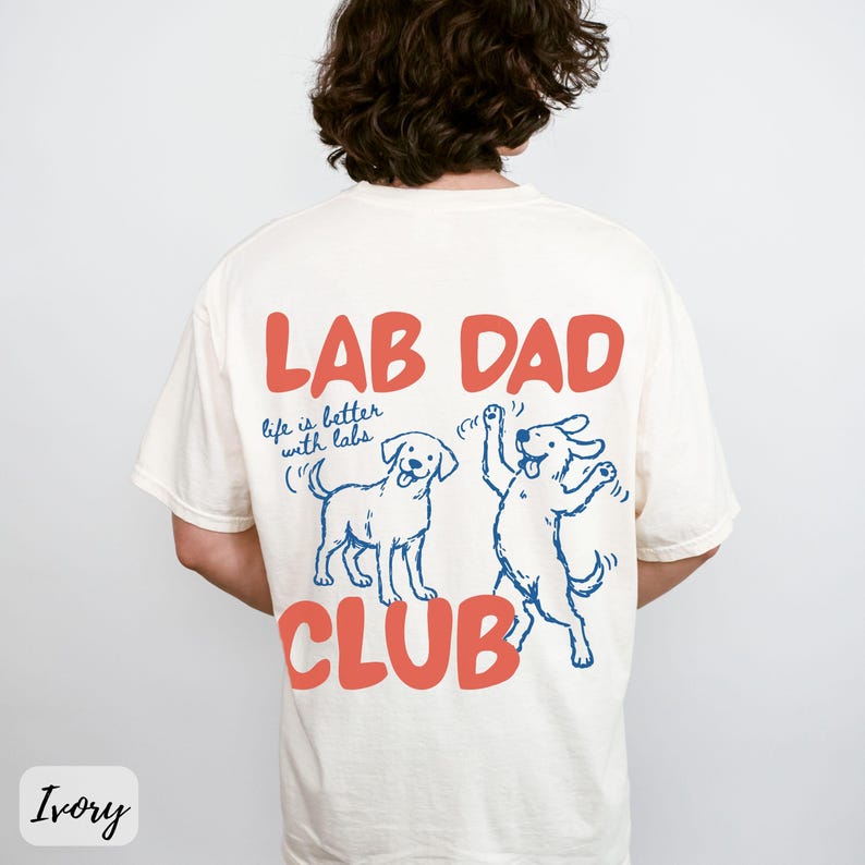 Puede incluir: Camiseta color marfil con las palabras "LAB DAD CLUB" en letras color coral. La camiseta presenta dos ilustraciones azules de l&iacute;nea de Labrador Retrievers y la frase "life is better with labs."