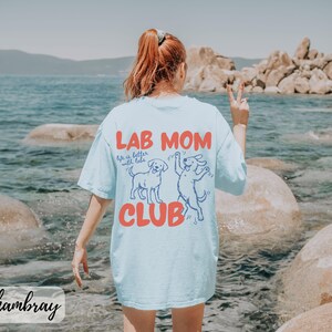 Puede incluir: Camiseta azul claro con la frase "LAB MOM CLUB" en rojo y un gr&aacute;fico de dos perros. La camiseta tambi&eacute;n tiene la frase "Life is better with labs". La camiseta est&aacute; hecha de un material suave.