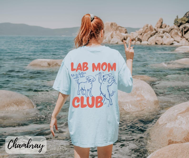 Puede incluir: Camiseta azul claro con la frase "LAB MOM CLUB" en rojo y un gr&aacute;fico de dos perros. La camiseta tambi&eacute;n tiene la frase "Life is better with labs". La camiseta est&aacute; hecha de un material suave.