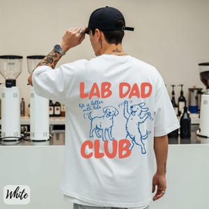 Puede incluir: Camiseta blanca con el texto "LAB DAD CLUB" en coral, con dos perros Labrador de dibujos animados en azul. Tambi&eacute;n incluye la frase "life is better with labs". La persona de la imagen lleva una gorra de b&eacute;isbol negra.