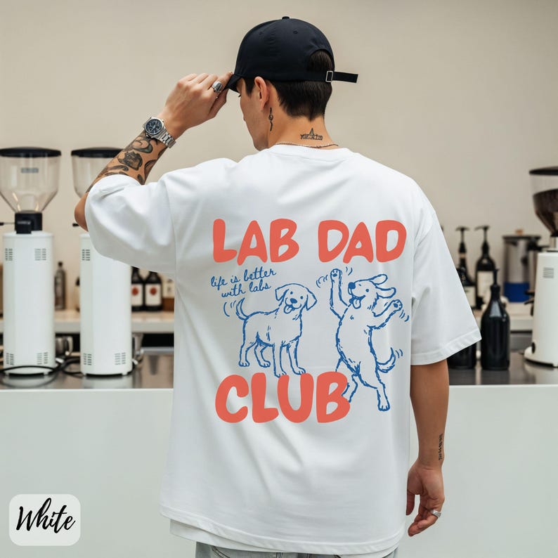 Puede incluir: Camiseta blanca con el texto "LAB DAD CLUB" en coral, con dos perros Labrador de dibujos animados en azul. Tambi&eacute;n incluye la frase "life is better with labs". La persona de la imagen lleva una gorra de b&eacute;isbol negra.