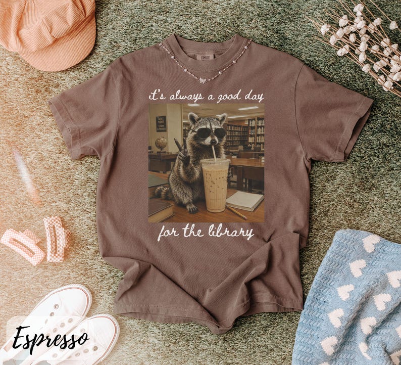 May include: A brown short-sleeve t-shirt featuring a graphic of a raccoon wearing sunglasses, making a peace sign, and holding an iced coffee. The text above the raccoon reads it's always a good day and below the raccoon reads for the library.