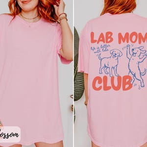 Puede incluir: Una camiseta rosa claro con el texto "LAB MOM CLUB" en coral y azul. La parte trasera de la camiseta presenta dos dibujos animados de perros labradores. La parte delantera de la camiseta tiene la palabra "Blossom" en cursiva.
