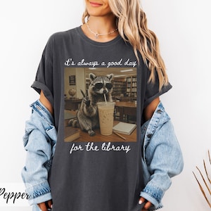 May include: A charcoal gray short-sleeve t-shirt featuring a graphic of a raccoon wearing sunglasses, holding a peace sign, and sitting next to an iced coffee. The text reads, "it's always a good day for the library" in white cursive script.