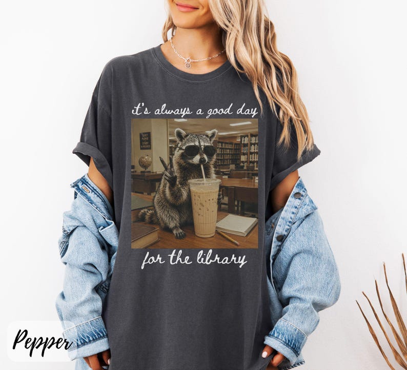 May include: A charcoal gray short-sleeve t-shirt featuring a graphic of a raccoon wearing sunglasses, holding a peace sign, and sitting next to an iced coffee. The text reads, "it's always a good day for the library" in white cursive script.