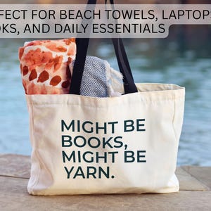 May include: A cream-colored tote bag with black handles, filled with items. The bag has the text "MIGHT BE BOOKS, MIGHT BE YARN" printed on it. The bag is filled with a patterned towel and other items.
