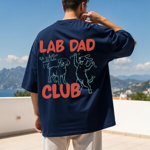 Puede incluir: Camiseta azul marino con el texto "LAB DAD CLUB" en naranja. La camiseta presenta dos ilustraciones de perros y la frase "life is better with labs". La camiseta tiene mangas cortas y un corte holgado.