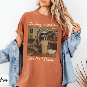 May include: A rust-colored t-shirt featuring a graphic of a raccoon wearing sunglasses, drinking an iced coffee, and making a peace sign in a library setting. The text above and below the image reads, it's always a good day for the library.