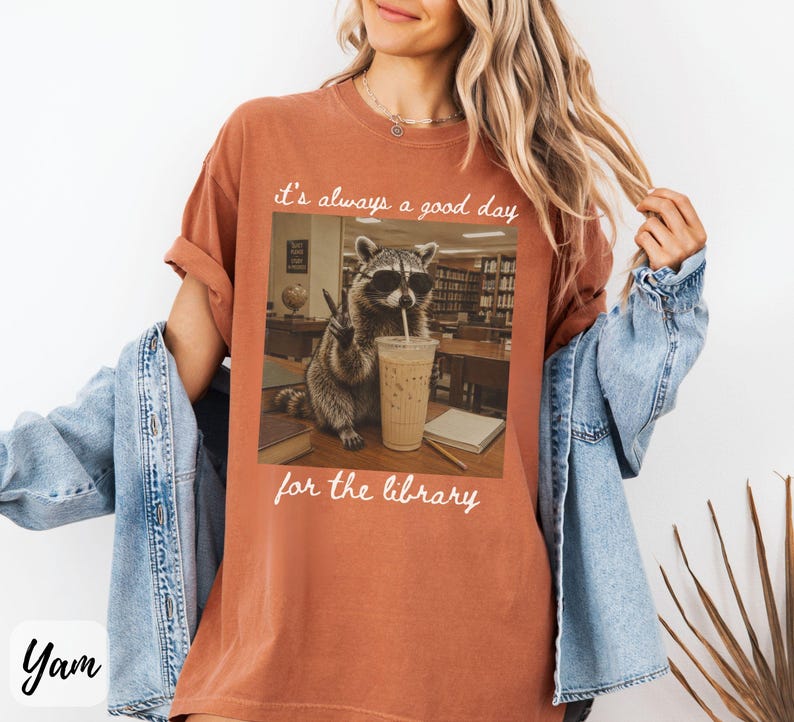 May include: A rust-colored t-shirt featuring a graphic of a raccoon wearing sunglasses, drinking an iced coffee, and making a peace sign in a library setting. The text above and below the image reads, it's always a good day for the library.