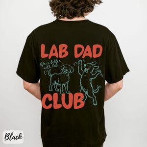 Puede incluir: Camiseta negra con el texto "LAB DAD CLUB" en rojo y la frase "life is better with labs" en blanco. Dos dibujos azules de perros Labrador Retriever. La camiseta tiene un corte holgado.