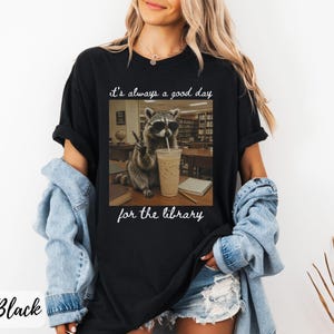 May include: A black t-shirt featuring a graphic of a raccoon wearing sunglasses, holding a drink with a straw, and making a peace sign. The text above the raccoon reads it's always a good day and below the raccoon reads for the library.