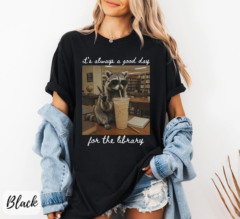 May include: A black t-shirt featuring a graphic of a raccoon wearing sunglasses, holding a drink with a straw, and making a peace sign. The text above the raccoon reads it's always a good day and below the raccoon reads for the library.
