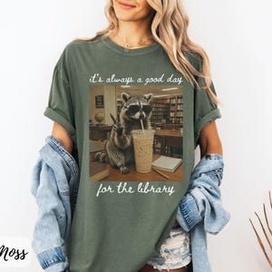 May include: A moss green short-sleeve t-shirt featuring a graphic of a raccoon wearing sunglasses, holding a drink, and making a peace sign in a library setting. The text above reads it's always a good day and below reads for the library.