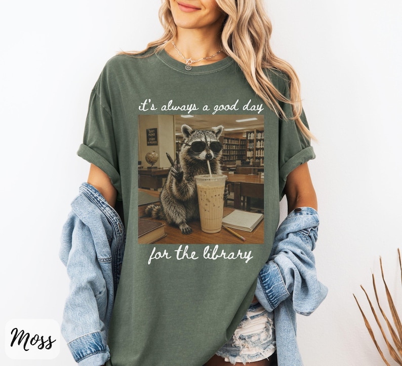 May include: A moss green short-sleeve t-shirt featuring a graphic of a raccoon wearing sunglasses, holding a drink, and making a peace sign in a library setting. The text above reads it's always a good day and below reads for the library.