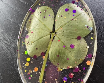 Op de afbeelding: Ovale sleutelhanger met een klavertjevier in heldere hars. Het klavertjevier is omgeven door kleurrijke glitter, waaronder paarse, gouden en iriserende spikkels. Een goudkleurige ketting en sleutelring zijn aan de bovenkant bevestigd.