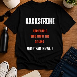Peut inclure: Un t-shirt noir à manches courtes avec un texte blanc et orange indiquant : BACKSTROKE FOR PEOPLE WHO TRUST THE CEILING MORE THAN THE WALL. Le vêtement présente une encolure ronde et un lettrage centré sur le devant du tissu noir uni.
