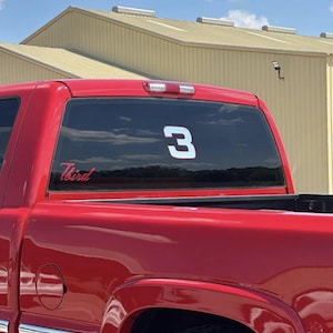May include: A red pickup truck with a large white number 3 on the rear window. The word "Third" is written in cursive below the window. The truck is parked outside on a sunny day.